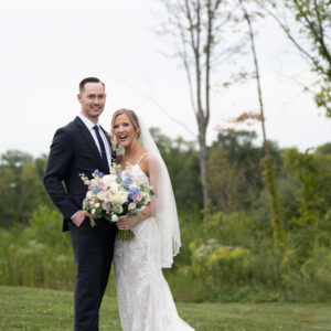 Bride and groom smiling on their wedding day outdoors. Bride holds a bouquet of flowers while the groom stands beside her in a suit against a scenic natural backdrop.
