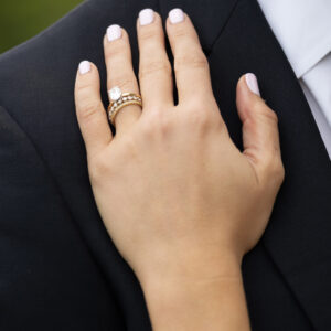 A woman's hand with elegant engagement and wedding rings rests on a man's shoulder, both well-dressed, symbolizing love and commitment on a special occasion.