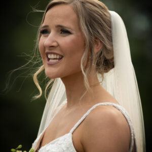 Bride in a white lace gown smiles joyfully, holding a bouquet of flowers on her wedding day. Her veil drapes elegantly, and a blurred natural background frames the scene.