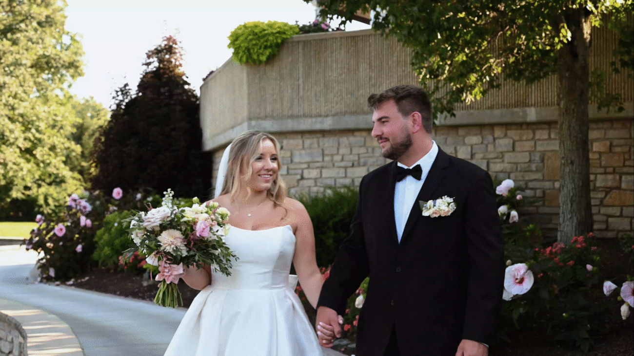 A happy couple walking hand in hand on their wedding day in an outdoor setting surrounded by greenery and flowers with the bride in a white dress and the groom in a black tuxedo.