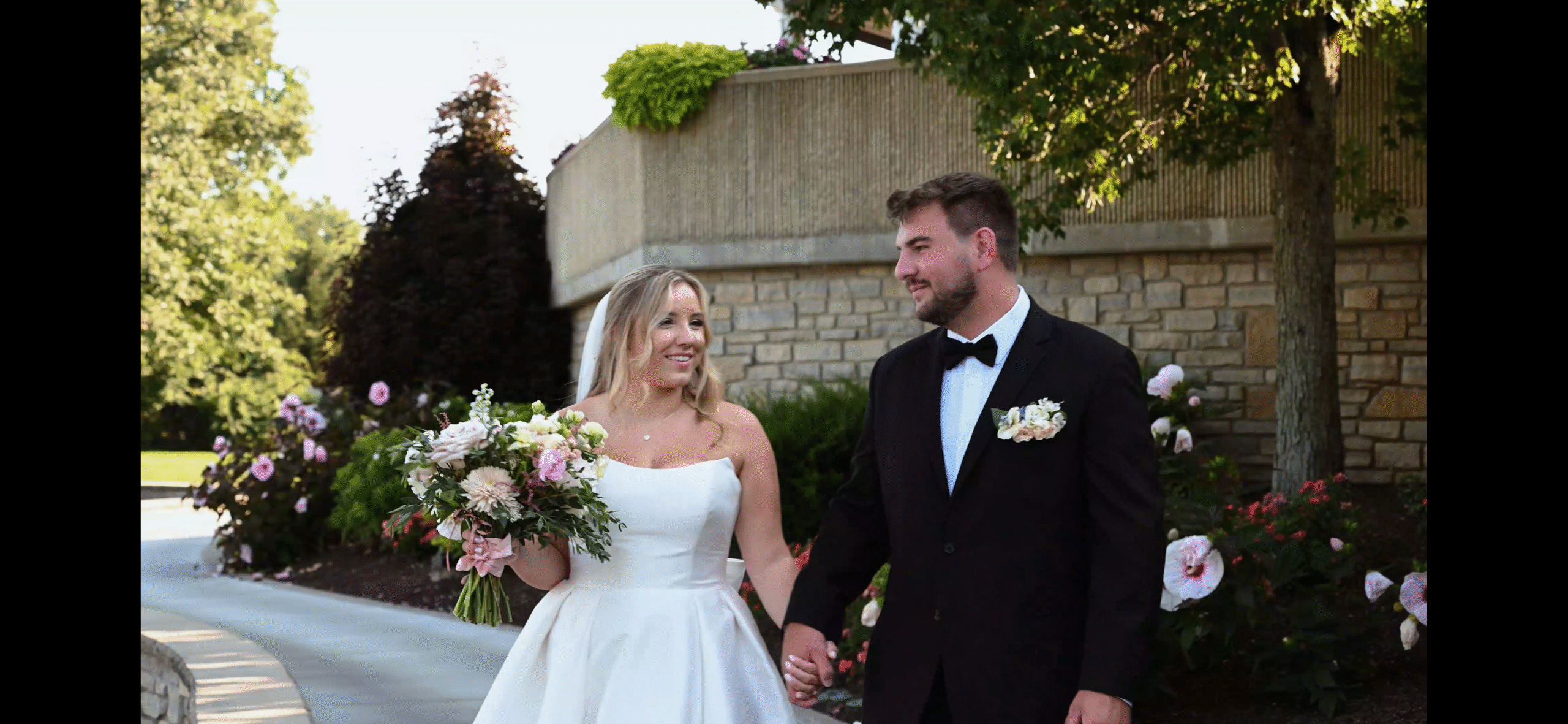 A happy couple walking hand in hand on their wedding day in an outdoor setting surrounded by greenery and flowers with the bride in a white dress and the groom in a black tuxedo.
