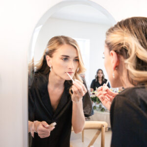 Woman applying lip gloss while looking in a mirror at a vanity table in a bright room reflecting a stylish and comfortable interior space enhancing her beauty routine.
