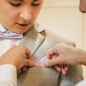 Young man in a beige suit getting a pink pocket square adjusted by another person. He is wearing a matching pink bow tie and a white shirt for a formal event.