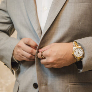 Man in gray suit with white shirt and lavender bow tie adjusts jacket buttons showcasing stylish watch and pocket square suitable for formal or business occasions