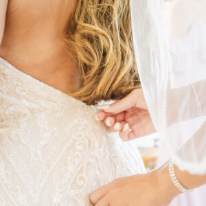 Bride in elegant white lace wedding dress with blonde hair being buttoned by bridesmaid, highlighting intricate gown details and special moments of the wedding day.