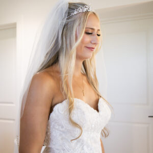 Elegant bride with long blonde hair smiles softly wearing a lace wedding dress and a sparkling tiara with veil in a serene indoor setting on her special day.