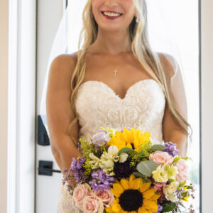 Bride in a white lace wedding gown holding a vibrant sunflower and rose bouquet. She smiles brightly with a background of a doorway filled with soft natural light.