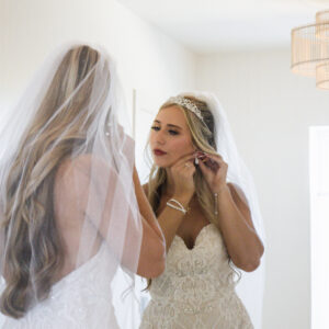Bride in elegant lace wedding gown and veil admires her reflection in a mirror while adjusting her earring in a softly lit room with stylish ceiling lamps