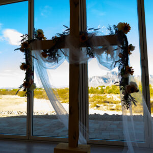 Wooden cross adorned with flowers and sheer fabric stands indoors against a large glass window overlooking a scenic outdoor landscape with a blue sky backdrop.