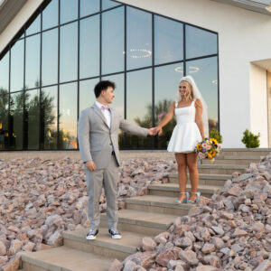 A couple in formal attire stands on stone steps in front of a modern glass building. The woman holds a bouquet and wears a white dress. The man is in a gray suit.