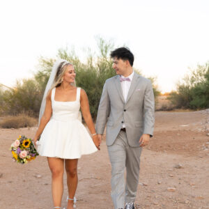 Bride in a short white dress and groom in a gray suit walking hand in hand on a dirt path surrounded by desert scenery with trees, carrying a bouquet of flowers.