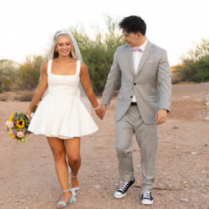Bride in a white dress and groom in a gray suit walking hand in hand on a desert path during sunset, with the bride holding a colorful bouquet.