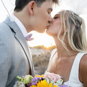 Bride and groom share a romantic kiss at sunset outdoors on their wedding day. Bride holds a vibrant bouquet with sunflowers while wearing an elegant white dress.