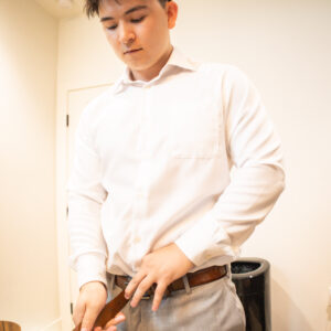 Young man adjusting a brown belt in a white dress shirt and gray trousers standing in a bright room with light walls conveying a sense of style and readiness.
