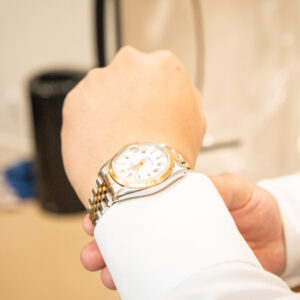 Close-up of a person's wrist wearing a classic gold and silver watch with a white dial. The watch is elegantly displayed against a white shirt sleeve in a well-lit room.