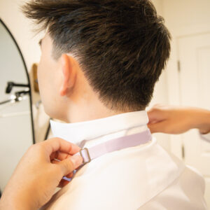 A person assists in adjusting a lavender bow tie on a man wearing a white dress shirt, preparing for a formal event in a brightly lit room.