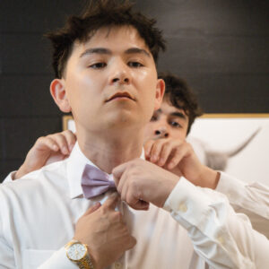 Young man in a white shirt adjusting his purple bow tie in front of a black wall. Another person assists him with the bow tie before a formal event or occasion.