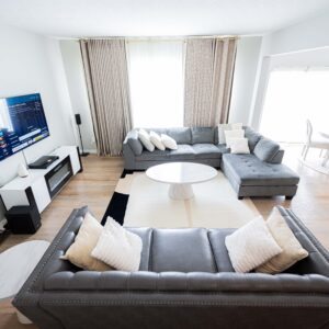 Bright modern living room with cozy gray sofas, a sleek white coffee table, wall-mounted TV, and large windows with elegant curtains. Hardwood floors and dining area.