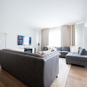 Modern living room with gray sectional sofas wooden flooring and a wall-mounted TV. Natural light filters through large windows providing a cozy and inviting atmosphere.