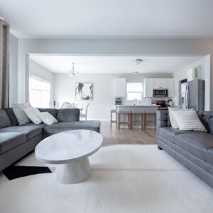 Modern open-plan living room with gray sofas and white marble coffee table leading into a bright kitchen featuring white cabinetry and stainless steel appliances.