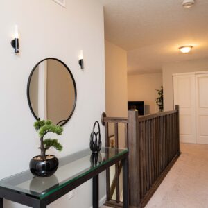 Modern hallway interior with a decorative mirror and a glass console table featuring a small potted plant. Soft lighting illuminates the beige carpeted floor and walls.