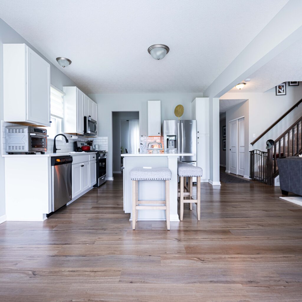 Modern open-plan kitchen and living room with white cabinets, stainless steel appliances, a cozy gray sofa, and wooden flooring providing a welcoming and spacious atmosphere.