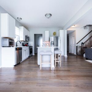 Modern open-plan kitchen and living room with white cabinets, stainless steel appliances, a cozy gray sofa, and wooden flooring providing a welcoming and spacious atmosphere.