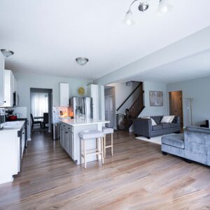 Bright modern open-concept living room with hardwood floors, featuring a cozy gray couch, flat-screen TV, and a kitchen with white cabinets and bar stools.