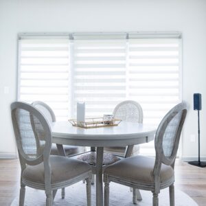 Bright and modern dining room with a round white table and four upholstered chairs set against large windows with blinds allowing natural light for a fresh and airy feel.