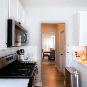 Bright modern kitchen with stainless steel appliances and white cabinets leading to a warmly lit dining area with wooden floors and elegant chairs around a table.