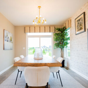 Bright dining room with a wooden table and white chairs, modern chandelier, artworks on the brick wall, and large windows offering a scenic outdoor view.