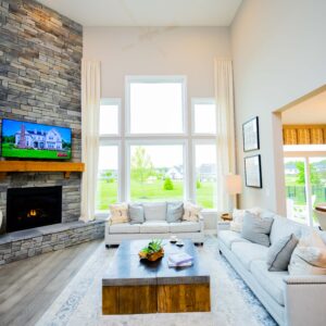 Modern living room with high ceiling and large windows, featuring a stone fireplace, TV, and comfortable sofas. Decor includes green plants and a wooden coffee table.
