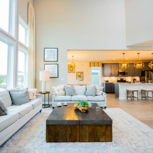 Spacious modern living room with large windows bright natural light gray sofas and a wooden coffee table open to a sleek kitchen with an island and pendant lighting.