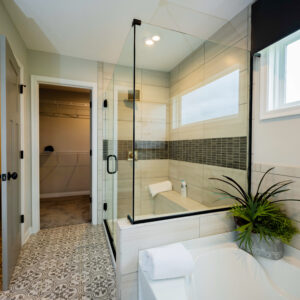 Modern bathroom with glass shower enclosure, tiled walls, and patterned floor. Features a mounted towel, potted plant, and natural light from a large window.