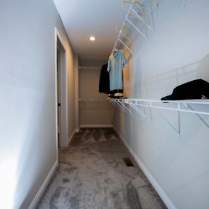 Empty walk-in closet with white walls and carpeted floor featuring a wall-mounted shelf with a few hanging clothes and a hat showcasing minimal storage space.