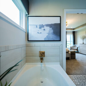 Modern bathroom interior featuring a white bathtub with a scenic wall art above. The room has natural light and adjoins a tastefully furnished bedroom through an open doorway.