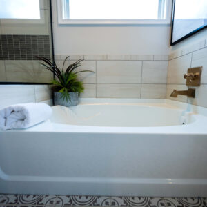 Modern bathroom with a white bathtub featuring rolled towels and a potted plant. The space is designed with neutral tiles and a stylish patterned floor for a sleek look.