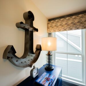 Cozy room with nautical decor featuring a large metal anchor wall art a stylish table lamp magazines and a window with patterned blinds for a seaside ambiance.