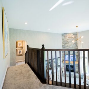 Modern hallway featuring carpeted flooring and stylish wooden railing overlooking a spacious living room with large windows and artistic wall decor enhancing the ambiance.