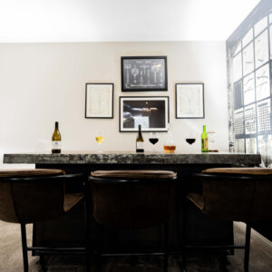 Modern home bar with stylish bar stools and a sleek countertop featuring wine bottles and glasses. Minimalist wall art enhances the contemporary design of the space.