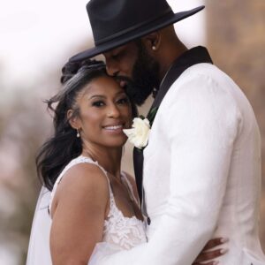 Bride and groom embracing on their wedding day. The groom is wearing a stylish hat and white suit while the bride is in a lace dress beaming with joy on a special occasion.