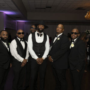 A group of six men dressed in formal black and white suits at a stylish indoor event with dim lighting and a chandelier in the background.