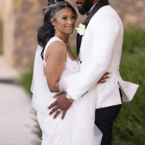 A joyful couple embraces on their wedding day with the groom in a stylish hat and the bride in an elegant gown against a natural stone background and lush greenery.