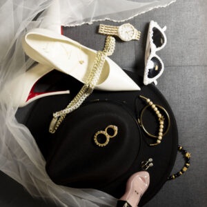 Elegant fashion accessories arranged on a gray surface including white high heels chic watches white sunglasses a black hat and various jewelry pieces for a stylish look