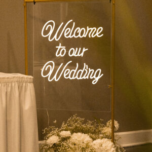 Welcome to our wedding sign with elegant white script displayed on a clear panel. Surrounded by a sophisticated arrangement of white flowers and set in a golden frame.