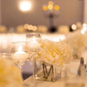 Elegant wedding table setting with glowing candlelight in glass holders and white floral centerpiece creating a romantic atmosphere. Soft focus and warm lighting enhance the mood.