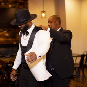 Man assisting another man in putting on a white suit jacket in a stylish wooden-themed room highlights classy fashion and elegance for a formal event or gathering.