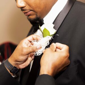 A person in a formal black tuxedo adjusting a white boutonniere on another man's lapel during a special occasion emphasizing elegance and attention to detail.