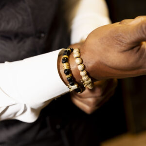Man in formal attire showcasing style with elegant black and gold beaded bracelets on wrist adding a touch of sophistication to the ensemble in a dimly lit setting.