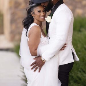 Elegant bride and groom embrace outdoors in stylish wedding attire. Bride wears a white gown and groom dons a white suit and black hat. Romantic setting with greenery.